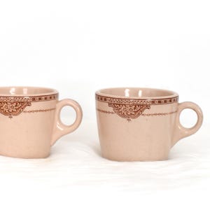 Peut inclure: Deux tasses à café en céramique rose clair avec un motif floral marron sur le bord. Chaque tasse a une anse arrondie. Les tasses sont sur une surface blanche.