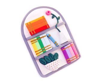 May include: A miniature bookshelf with three shelves, each filled with colorful books. The top shelf has a stack of red, orange, and yellow books. The middle shelf has a stack of green, blue, and yellow books. The bottom shelf has a stack of pink, purple, and blue books. There is a small green plant in a white pot with red polka dots on the bottom shelf.