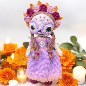 May include: A handmade crochet doll in the style of a Day of the Dead figure. The doll has a purple dress, a floral crown, and braided brown hair. The doll's face is painted with a skull design. The doll is surrounded by orange flowers and lit candles.