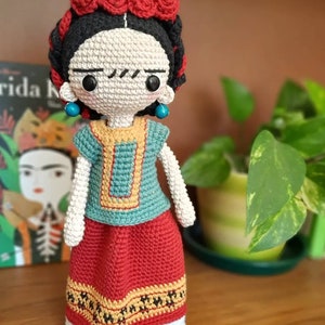 May include: Crocheted doll with a red and white dress, black hair, and a flower crown. The doll is wearing turquoise earrings and has a painted-on mustache.