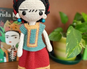 May include: Crocheted doll with a red and white dress, black hair, and a flower crown. The doll is wearing turquoise earrings and has a painted-on mustache.