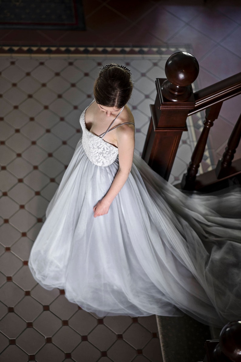May include: A woman wearing a light gray tulle wedding dress with a lace bodice. She is standing on a staircase with a wooden railing.