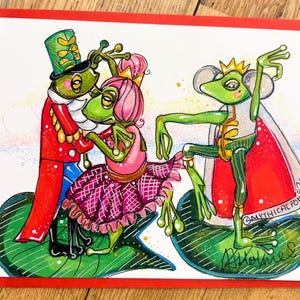 May include: A whimsical greeting card featuring three anthropomorphic frogs in a fairytale setting. One frog wears a red and blue uniform, another a pink dress, and the third a regal cape. The artwork is set on a white background with a red border.