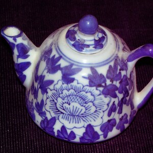May include: A small, white and blue ceramic teapot with a floral design. The teapot features a rounded body, a curved spout, and a handle. The lid has a small knob. The floral pattern includes a large flower and leaf accents.