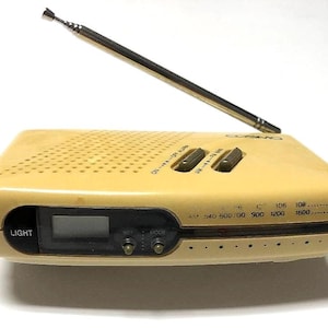 May include: A beige portable radio with a telescoping metal antenna, a digital display, and a speaker grille on top. The device features a black control panel labeled LIGHT, SET, and MODE, with AM and FM frequency markings printed on the front.