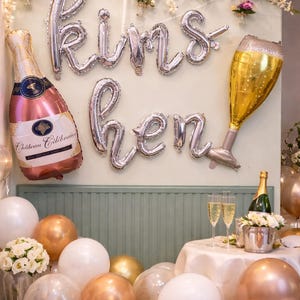 Könnte beinhalten: Festliche Dekoration mit silbernen Luftballons, die "Kims hen" bilden, und Champagner-Themen. Enthält einen Luftballon in Form einer Champagnerflasche, einen Luftballon in Form eines Sektglases und verschiedene goldene, weiße und transparente Luftballons.