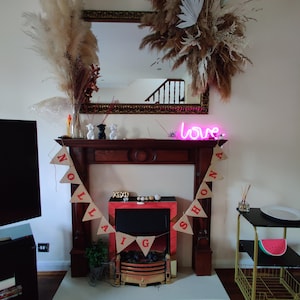 May include: A fireplace mantel decorated with dried pampas grass, a mirror, and a pink neon "love" sign. A banner with red letters spells out a message. A small table displays a watermelon decoration.