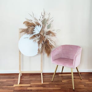 Op de afbeelding: Een lichtroze fluwelen fauteuil met gouden poten staat naast een houten bord versierd met gedroogde bloemen en een lichtblauw cirkelvormig element. De opstelling bevat pampasgras en ander gedroogd blad.