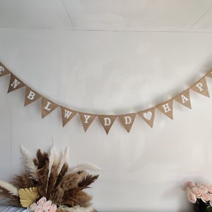 Puede incluir: Una pancarta de arpillera con las palabras "Pennlydd Hapus" y un corazón en medio. La pancarta está colgada en una pared blanca.