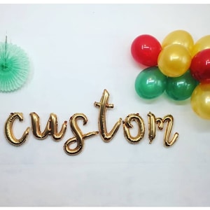 May include: Gold foil letter balloons spelling out the word "custom" against a white background. A green and gold balloon garland is in the top right corner of the image.