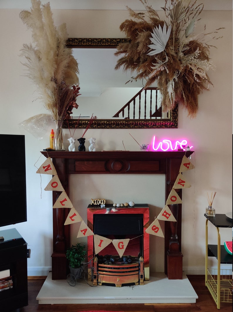 May include: A decorated fireplace with a mirror, dried floral arrangements, and a banner that reads "NOLA & SHONA". A neon sign displays the word "love". Decorative items include small figurines and a miniature bicycle.