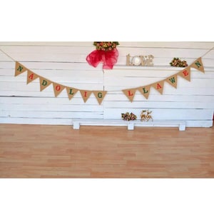 May include: A festive banner with the words "NADOLIG LLAWEN" in red and green letters, strung across a white wooden backdrop. The banner is made of burlap triangles. A red bow and floral arrangement are visible above.