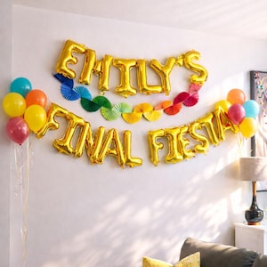 May include: A wall decorated with gold foil balloons spelling "EMILY'S FINAL FIESTA", alongside colourful paper fans and balloons in yellow, orange, blue, and red. A celebratory party decoration.