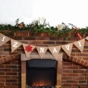 May include: A burlap banner with the words "FELIZ NAVIDAD" in white letters hangs above a brick fireplace. A red snowflake adorns the center pennant. The fireplace is decorated with greenery and candles, creating a festive holiday scene.
