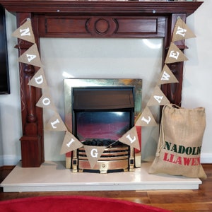May include: A burlap banner with white letters spelling "NADOLIG LLAWEN" hangs above a fireplace. A matching burlap sack with the same text in red and green sits to the right. The fireplace has a gold frame.