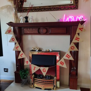 May include: A wooden fireplace mantel with a mirror and a neon "love" sign. A banner with red letters spelling "Nollaig Shona" hangs in front of the fireplace. Decorative items include vases, figurines, and a small bicycle planter.