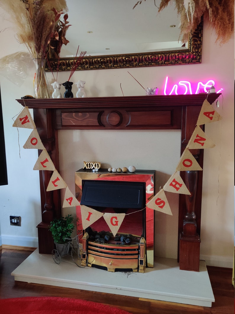 May include: A wooden fireplace mantel with a mirror and a neon "love" sign. A banner with red letters spelling "Nollaig Shona" hangs in front of the fireplace. Decorative items include vases, figurines, and a small bicycle planter.