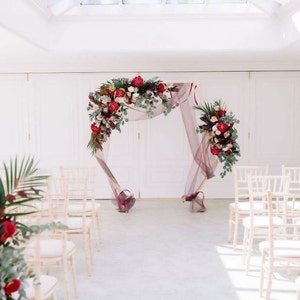 May include: A wedding ceremony setup with white chairs and a floral arch decorated with red and pink flowers. The arch is made of a sheer fabric and is draped with greenery.