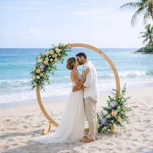 May include: A beach wedding ceremony with a wooden circular arch decorated with yellow and purple flowers. A couple kisses under the arch on a sandy beach with the ocean in the background.