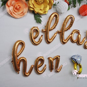 Könnte beinhalten: Goldene Folien-Buchstabenballons, die "ellas hen" buchstabieren, mit einem goldenen und weißen Diamant-Ringballon.