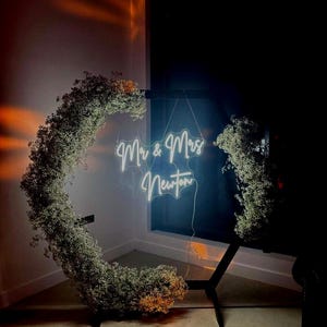 May include: A neon sign with the words "Mr & Mrs Newton" hangs in front of a dark background. The sign is surrounded by a circular floral arrangement of white flowers. The sign is supported by a black metal stand.