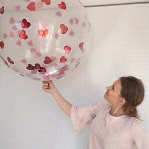 Può includere: Un grande palloncino trasparente pieno di coriandoli a forma di cuore rossi. Il palloncino è tenuto da una persona che indossa un top rosa. Il palloncino è rotondo e sembra gonfiato. I coriandoli sono sparsi nel palloncino.