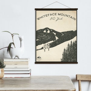 Puede incluir: Una ilustración de estilo vintage de la montaña Whiteface en Nueva York. La ilustración presenta una representación en blanco y negro de la montaña con una pista de esquí y árboles. El texto "Whiteface Mountain New York" está impreso en negro en la parte superior de la ilustración.