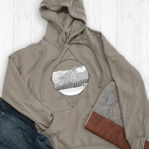 Könnte beinhalten: Ein hellbraunes Kapuzenpullover mit einem Grafikdruck eines Gebirges in einem Kreis und einem Dreieck. Der Text "Copper Mountain" ist unter dem Grafikdruck gedruckt.