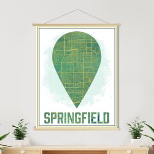 Pode incluir: Um mapa verde e amarelo de Springfield, Missouri, em forma de alfinete, sobre um fundo branco com um efeito de aquarela azul claro. O texto "SPRINGFIELD" está abaixo do mapa.