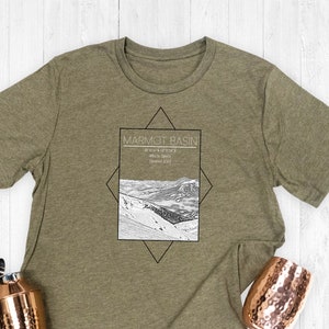 May include: Olive green t-shirt featuring a black and white graphic of a mountain range with the text "MARMOT BASIN". The shirt is made of a soft, comfortable material.