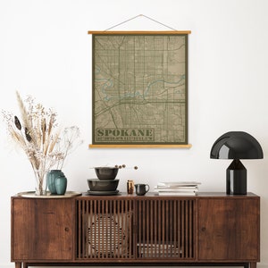 May include: A beige canvas wall hanging with a map of Spokane, Washington. The map is printed in green and blue on a beige background. The text "SPOKANE 47.63926.0°N 117.2511.1°W" is printed in green at the bottom of the map.