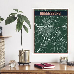 May include: A dark green map print with white lines showing the streets of Greensburg. The map is framed with a white border and a thin, copper-coloured border. The print is hanging on a white wall.