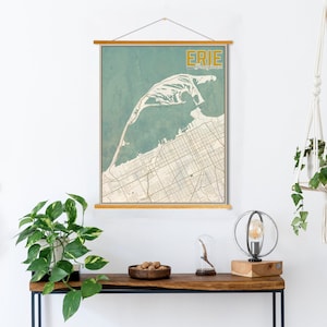 May include: A vintage-style map print of Erie, Pennsylvania, with a light blue background and brown lines depicting streets. The map is framed with a light brown wooden frame and hanging on a white wall.