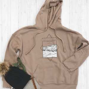 May include: A tan hoodie with a black and white graphic print of a mountain scene. The graphic includes the text "St. Anton am Arlberg" and "Vorarlberg, Austria".