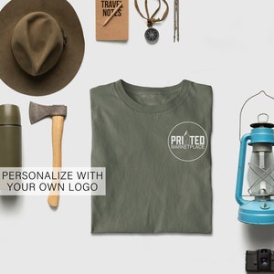 May include: An olive green t-shirt with a white circular logo that says "PRINTED MARKETPLACE". The image also includes a brown hat, an axe, a compass, a lantern, and a camera. The text "PERSONALIZE WITH YOUR OWN LOGO" is also visible.