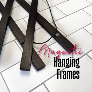 Op de afbeelding: Zwarte magnetische hangende frames van hout met een zwart touwtje eraan. De frames liggen op een witte betegelde ondergrond. De tekst "Magnetische hangende frames" staat in roze letters onder de frames.