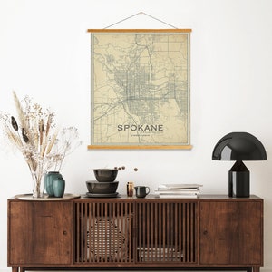 May include: A vintage-style map print of Spokane, Washington, hanging on a white wall above a wooden dresser with a black lamp and decorative bowls. The map is printed on cream-colored paper with blue lines and text.