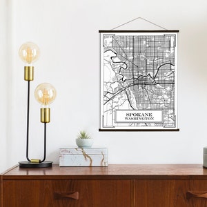 May include: Black and white map print of Spokane, Washington, with the city name and state printed at the bottom. The map is hanging on a wall with a wooden dowel rod at the top and bottom.