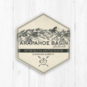 May include: A vintage-style illustration of a mountain range with the text "ARAPAHOE BASIN Colorado" and coordinates "39°38'33.1"N 105°52'19.0"W". The text "ELEVATION 13,050 FT" and "EST 1946" are also included.