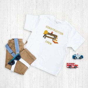 K&ouml;nnte beinhalten: Wei&szlig;es T-Shirt mit einem Bauarbeiter-Motiv. Das Motiv zeigt einen gelben Helm, eine schwarz-gelbe Baustellenabsperrung und einen orangefarbenen Verkehrskegel. Der Text "CONSTRUCTION CREW" ist in Gelb auf dem Shirt gedruckt.