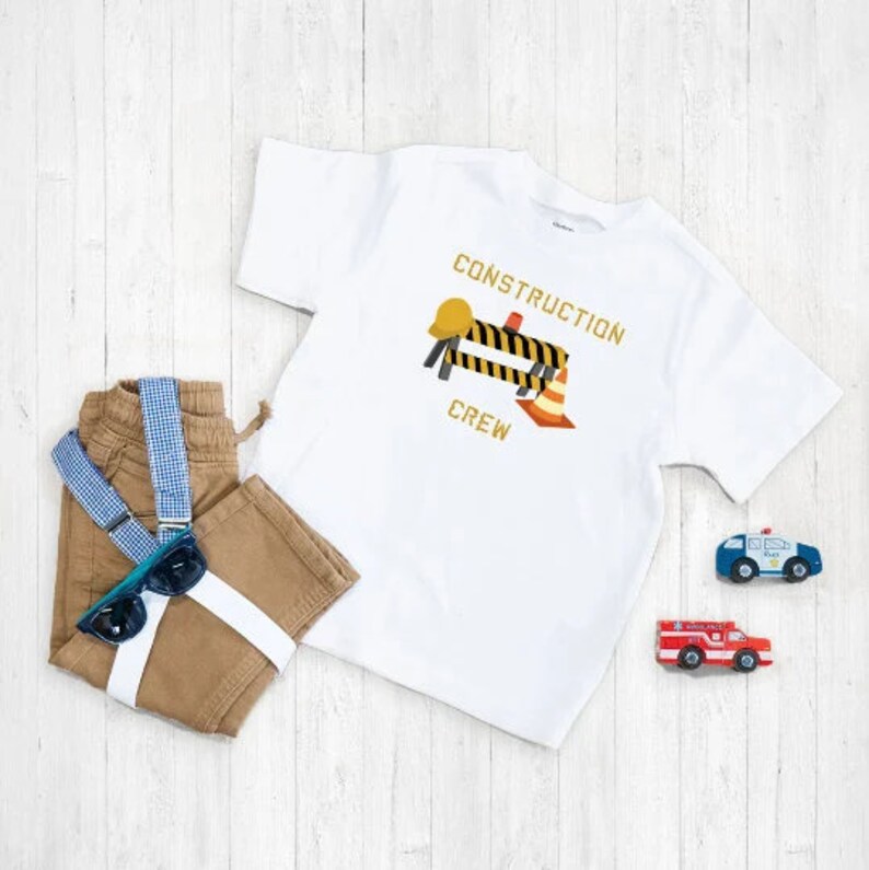 K&ouml;nnte beinhalten: Wei&szlig;es T-Shirt mit einem Bauarbeiter-Motiv. Das Motiv zeigt einen gelben Helm, eine schwarz-gelbe Baustellenabsperrung und einen orangefarbenen Verkehrskegel. Der Text "CONSTRUCTION CREW" ist in Gelb auf dem Shirt gedruckt.