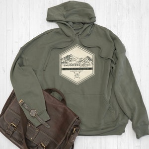 Könnte beinhalten: Ein grüner Kapuzenpullover mit einem weißen sechseckigen Patch, der eine Bergkette zeigt und den Text "Smugglers' Notch" mit Koordinaten und Höhe.