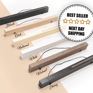 May include: Six wooden poster hangers in different colours: black, oak, natural, white, walnut, and ebony. Each hanger has a black string attached to it and the words "www.printedmarketplace.com" are printed on the bottom of each hanger. A white circle with gold text says "BEST SELLER" with five gold stars and "NEXT DAY SHIPPING".