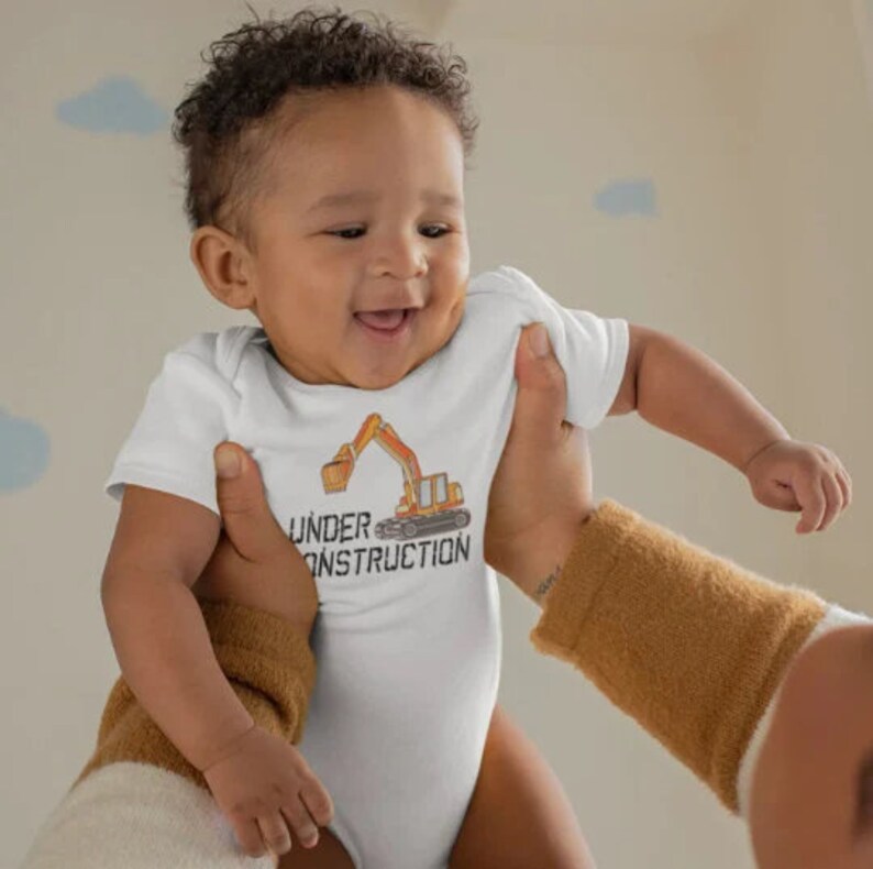 K&ouml;nnte beinhalten: Ein wei&szlig;er Baby-Strampler mit einem gelben Baufahrzeug-Motiv und dem Text "UNDER CONSTRUCTION".