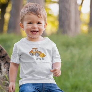 K&ouml;nnte beinhalten: Ein kleines Kind tr&auml;gt ein wei&szlig;es T-Shirt mit einem Comic-Bagger und dem Text "I DIG IT". Das Kind l&auml;chelt und tr&auml;gt eine blaue Jeans.