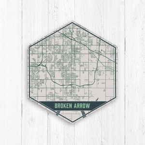 Puede incluir: Un mapa hexagonal de Broken Arrow, Oklahoma, en verde y blanco. El mapa muestra carreteras y edificios, con el nombre de la ciudad en una pancarta azul oscuro en la parte inferior. El mapa está sobre un fondo de madera blanca.