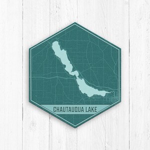 May include: A teal blue hexagonal sign with a map of Chautauqua Lake in New York. The lake is outlined in a lighter blue color. The sign has a distressed, vintage look and the text "Chautauqua Lake" is printed in white.