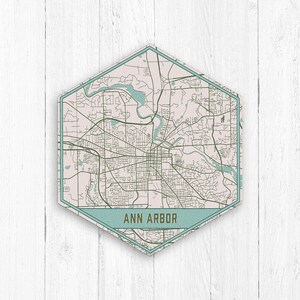 May include: A hexagonal map of Ann Arbor, Michigan, with a light pink background and dark green lines representing streets. The city name is written in a dark green font on a light blue banner at the bottom of the map.