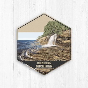May include: A hexagonal vintage travel poster with a scenic image of a waterfall cascading into a rocky shoreline. The poster has a tan background with a black border and the text "MUNISING MICHIGAN" at the bottom.
