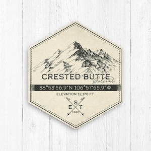 May include: A vintage-style illustration of a mountain range with the text "CRESTED BUTTE Colorado" and coordinates "38°53'56.9"N 106°57'55.9"W". The text "ELEVATION 12,170 FT" and "EST 1880" are also included.